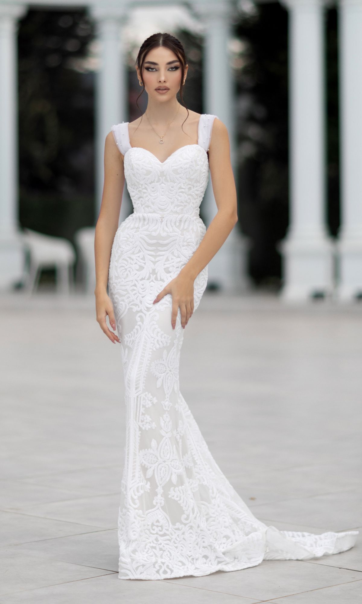 A woman stands outdoors before white columns wearing the Tina Holly TW103, an off-shoulder, floor-length white bridal gown.